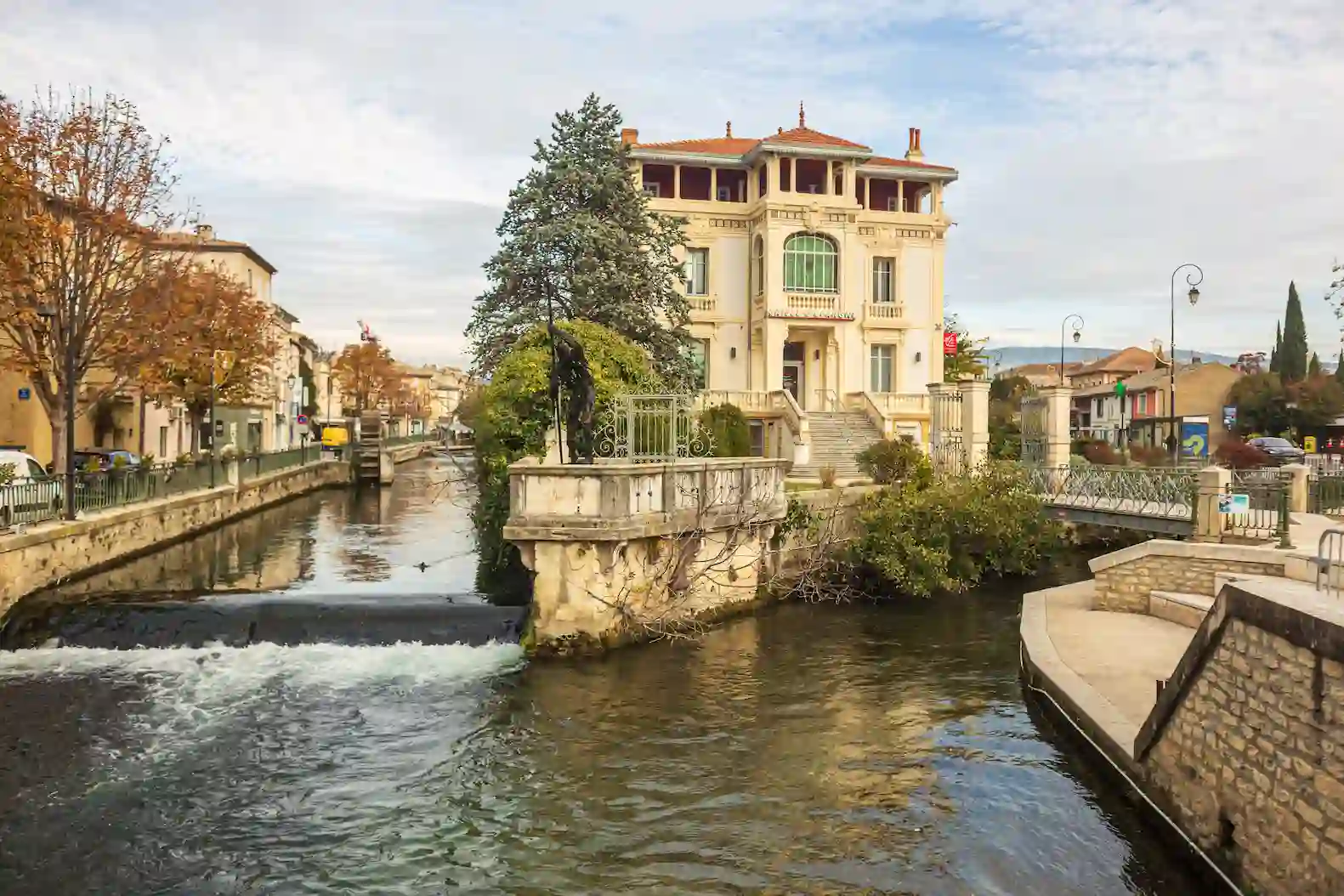 Vue des canaux de L’Isle-sur-la-Sorgue avec une villa bourgeoise et un pont en pierre, typiques de la Venise provençale.