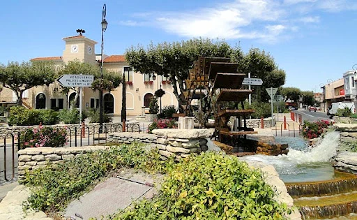 Vue de la place à Noves, avec sa fontaine et ses aménagements paysagers, capturant l’essence d’un village provençal typique.